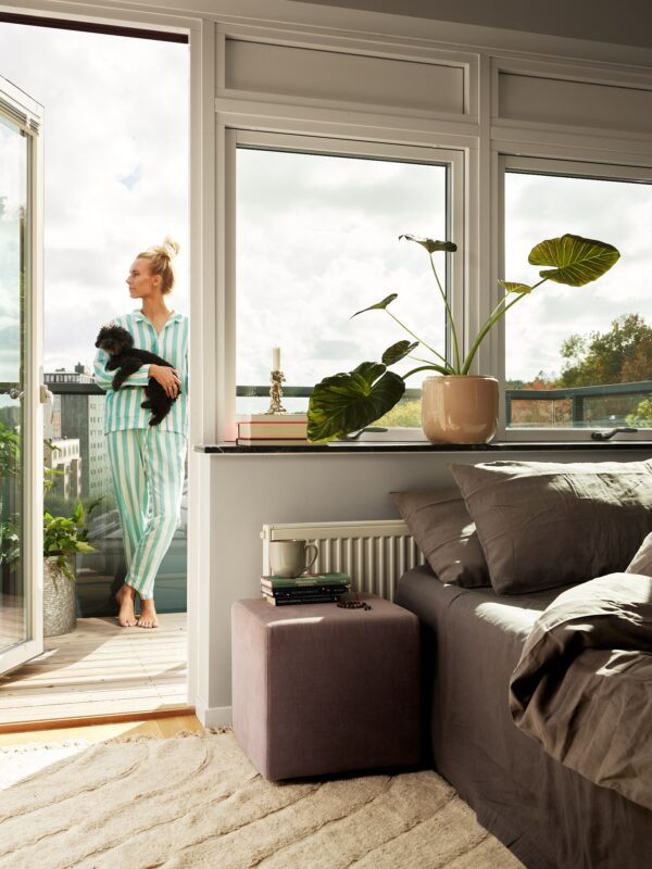 En kvinna i randig pyjamas på en balkong utanför ett sovrum håller en liten svart hund i famnen